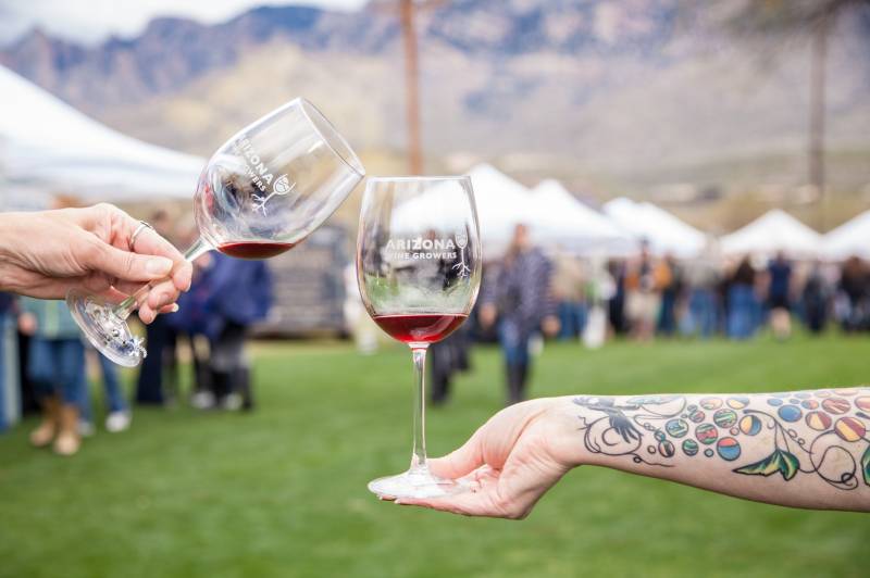 Two hands hold wine glasses in the air appearing to cheers in front of a crowd of people at Sedona WineFest.